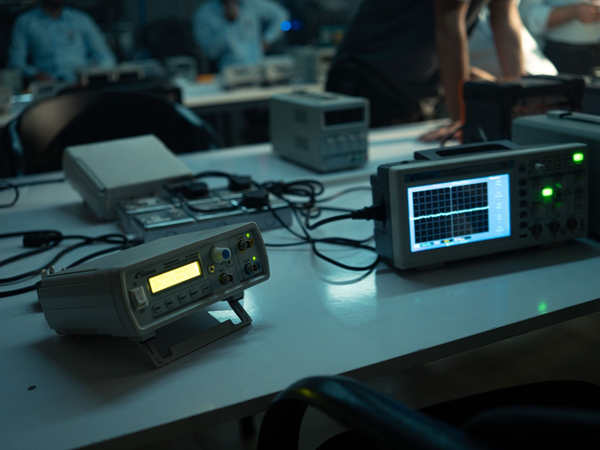 A dim electronics lab bench with an oscilloscope, a signal generator and a bench supply, people working out of focus behind. Illustrative photograph.